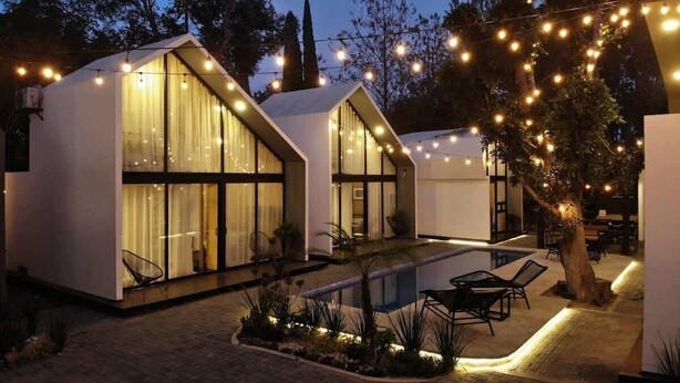 A modern outdoor pool area with two glass-enclosed structures, string lights, and a tree.