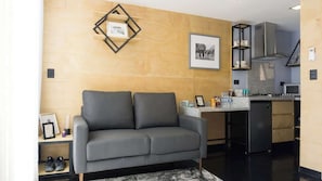 Un salon moderne avec un canapé gris, une petite table basse et un coin cuisine équipé d’une cuisinière et de placards.