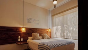 A bedroom with a wooden headboard, a bedside table with a lamp, a bed with a striped blanket, and a window with blinds. - Comfortable Apartment with Pool & Outdoor Oasis (Puerto Escondido)