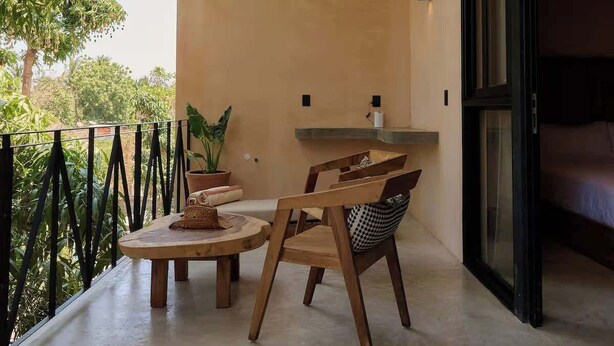 Un balcone con tetto di paglia, un tavolo di legno rotondo, due sedie, una pianta in vaso e un letto visibile attraverso una porta aperta.
