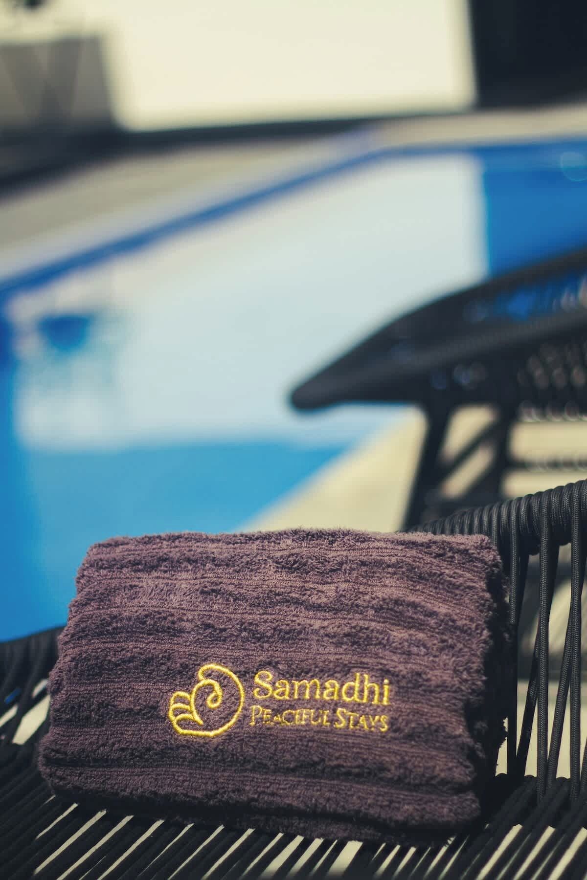 Uma toalha com a inscrição 'Samadhi Peaceful Stays' bordada, colocada sobre uma cadeira perto de uma piscina.
