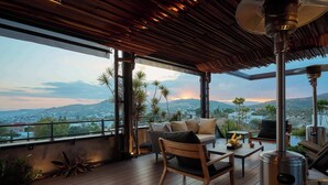 Une terrasse en bois avec un canapé, des chaises et une table, offrant une vue sur le paysage urbain et les montagnes.