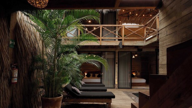 Une terrasse en bois avec des chaises longues, une grande plante en pot et un balcon doté d’un toit de chaume.