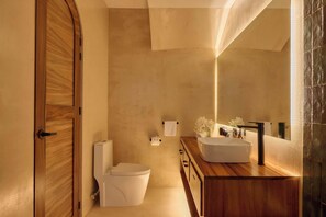 A modern bathroom with a wooden door, a white toilet, a wooden vanity with a sink, and a mirror. - Oceanview King Suite w/ Soaking Tub & Rooftop Terrace (Puerto Escondido)