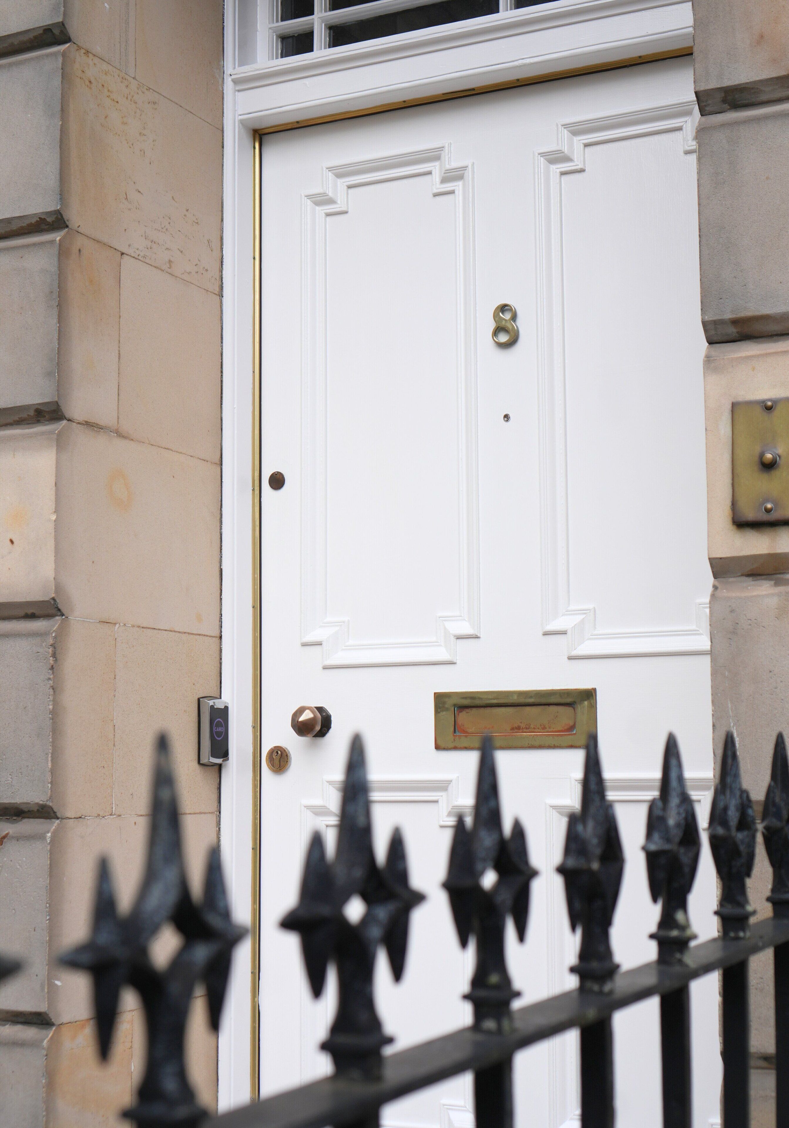 A white door with a brass knocker and a small plaque, set in a stone wall with a metal gate in the foreground.
