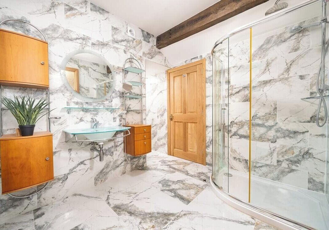 Un baño con pared de mármol, puerta de madera, cabina de ducha de vidrio, lavabo con pila de vidrio y un espejo.