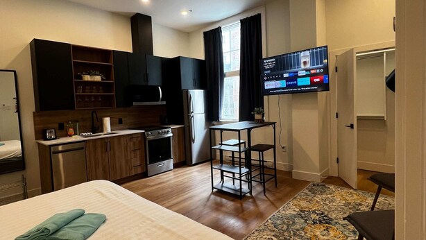 Moderni hotellihuone, jossa on sänky, pieni keittonurkkaus, ruokailutila ja televisio, josta voi katsoa suoratoistopalveluita.
