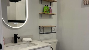 Moderni kylpyhuone, jossa on valkoinen pesuallas, musta hana, wc-istuin ja peili, jonka kehys on valaistu. WC-istuimen yläpuolella on kaksi puuhyllyä, joista toisella on hygieniatuotteita ja toisella koriste-esine.