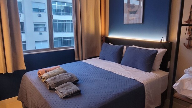 Hotellihuone, jossa on suuri sänky, yöpöytä, työpöytä, tuoli, ikkuna, jossa on verhot, ja seinälle kiinnitetty taideteos.