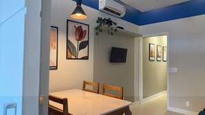 Un espace repas comprenant une table, des chaises, une suspension, un téléviseur fixé au mur et des œuvres d’art encadrées.