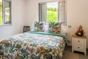 A bedroom with a bed, two windows with shutters, a bedside table with a vase, and a green bedspread. - PARADISE II (Bora Bora)