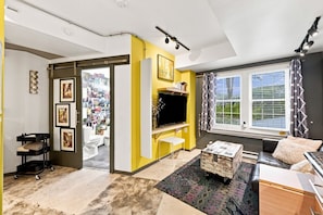 A living room with a television, a sofa, a coffee table, and a window with curtains. - The Steele Mill + Loft (Indianapolis)