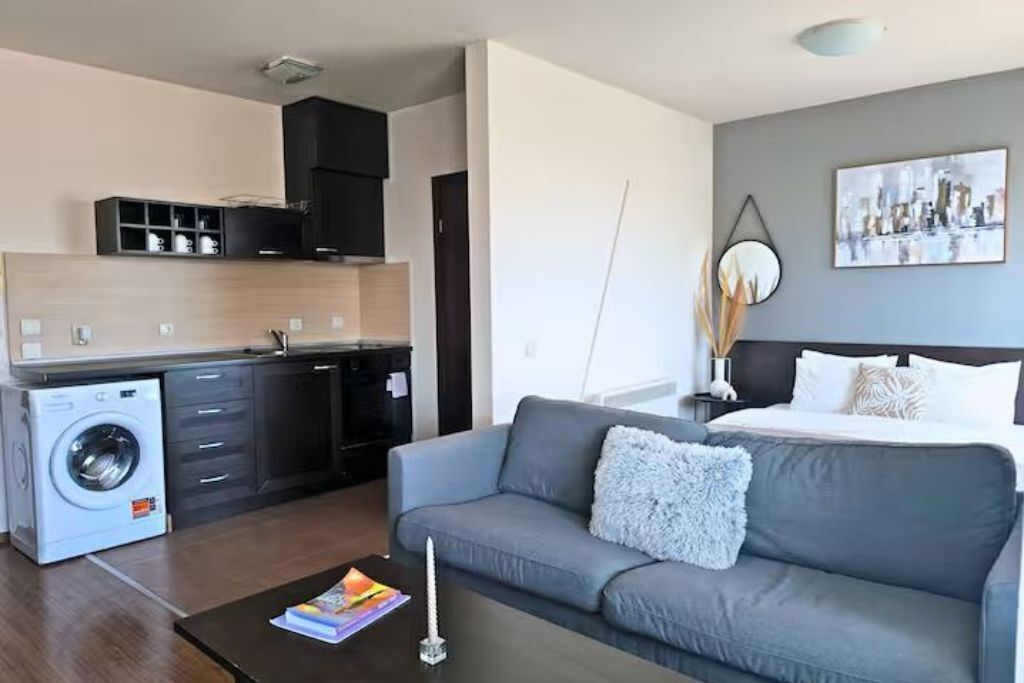 Een functionele woonruimte met een bank, een klein tafeltje, een wasmachine, een kitchenette en een bed met hoofdeinde.