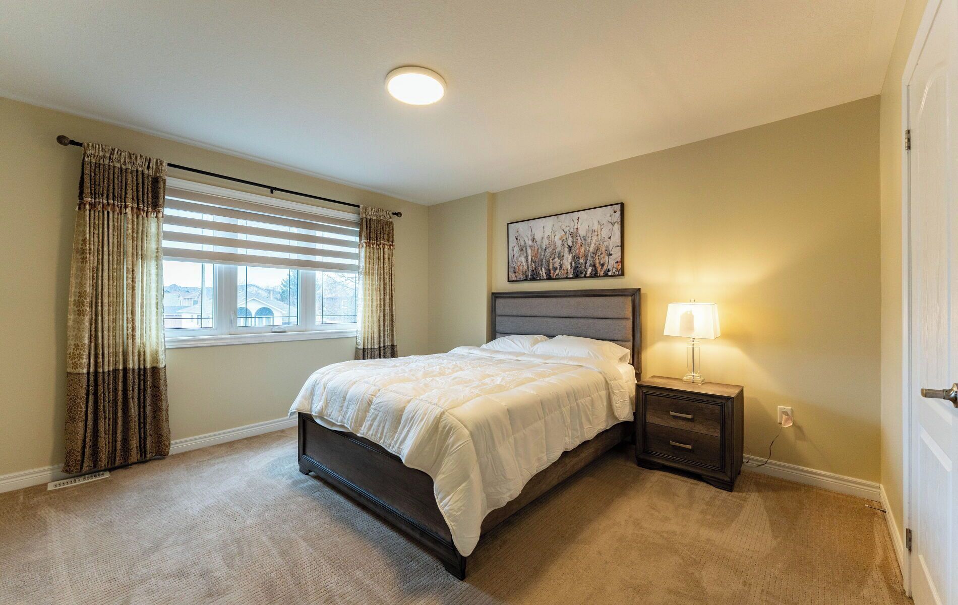 A bedroom with a bed, a nightstand with a lamp, a window with blinds, and a painting on the wall.