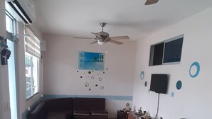 A living room with a sectional sofa, a ceiling fan, a flat-screen TV, and a window with blinds.