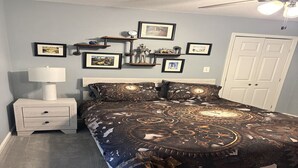 Quarto com cama, mesa de cabeceira, abajur e prateleiras na parede com quadros.