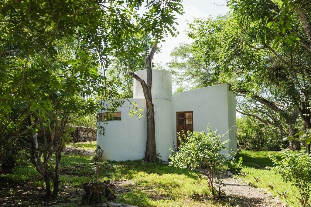 A small, white building with a single window and a door, surrounded by greenery and trees.