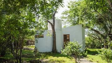 A small, white building with a single window and a door, surrounded by greenery and trees.