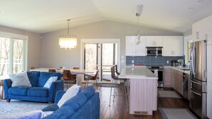 Eine moderne Küche mit Edelstahlgeräten, ein Essbereich mit Tisch und Stühlen sowie ein Wohnzimmer mit blauem Sofa.