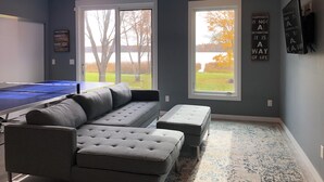 Ein Zimmer mit einem Tischtennis-Tisch, einem grauen Ecksofa, zwei Hocker und großen Fenstern mit Blick auf Bäume und einen Wasserfall.