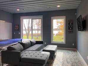 Ein Zimmer mit einem Tischtennis-Tisch, einem grauen Ecksofa, zwei Hocker und großen Fenstern mit Blick auf Bäume und einen Wasserfall.