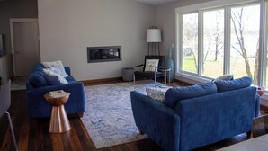 Ein modernes Wohnzimmer mit zwei blauen Sofas, einem Couchtisch, einer Stehlampe und einem großen Fenster mit Blick ins Freie.