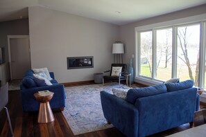 Ein modernes Wohnzimmer mit zwei blauen Sofas, einem Couchtisch, einer Stehlampe und einem großen Fenster mit Blick ins Freie.
