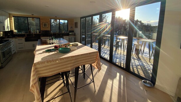 A modern kitchen with a large island, a dining area with a table, and a view of a snowy deck. - Mollusk Alaska Retreat, Private Coastal Escape in Homer, AK (Homer)