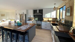 A modern kitchen with white cabinets, a central island with stools, a built-in oven, and a large window providing natural light. - Mollusk Alaska Retreat, Private Coastal Escape in Homer, AK (Homer)