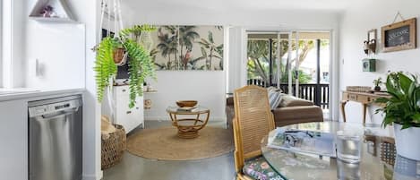 Una cocina moderna con lavavajillas, un comedor con mesa de vidrio y sillas de mimbre, y un espacio de sala con sofá y vistas al exterior.