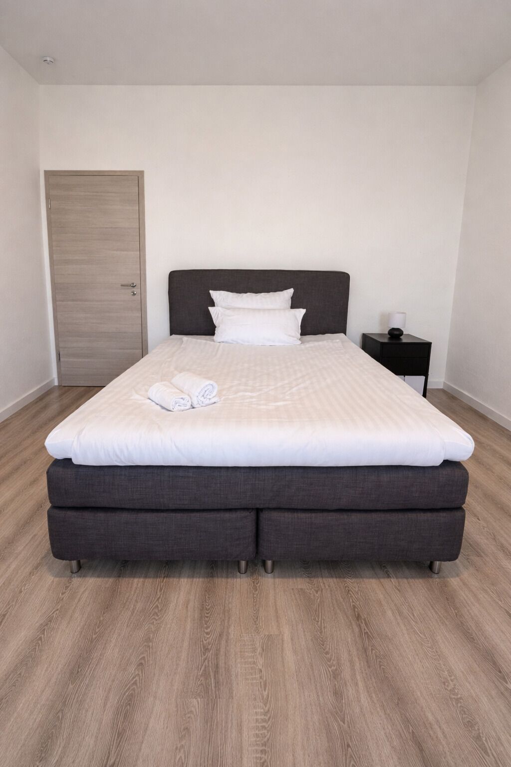 A bedroom with a bed, a nightstand, a wooden door, and a light switch on the wall.