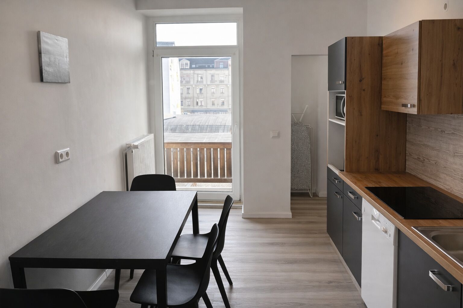 Une cuisine moderne avec un plan de travail en bois, une table à manger noire avec des chaises, et une vue sur un immeuble par la fenêtre.
