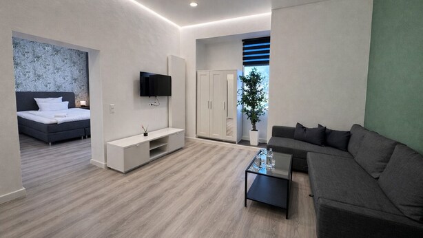 A modern living room with a sofa, a coffee table, a TV mounted on the wall, a bed with a headboard, and a potted plant. - MIILOX Apartments Chemnitz (Chemnitz)