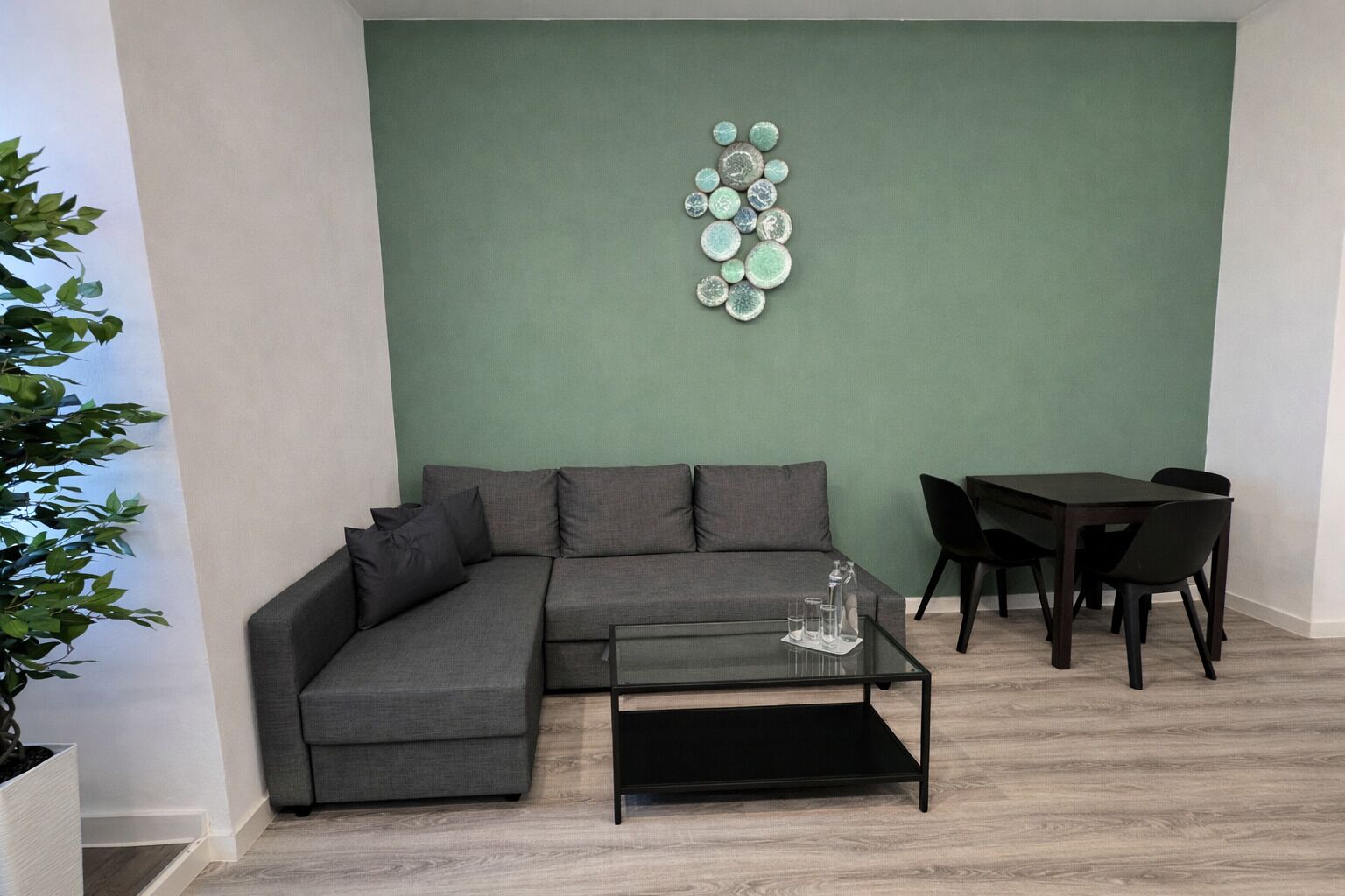 Una sala de estar con un sofá seccional gris, una mesa de centro de vidrio, una mesa de comedor con sillas y una pared de acento verde.
