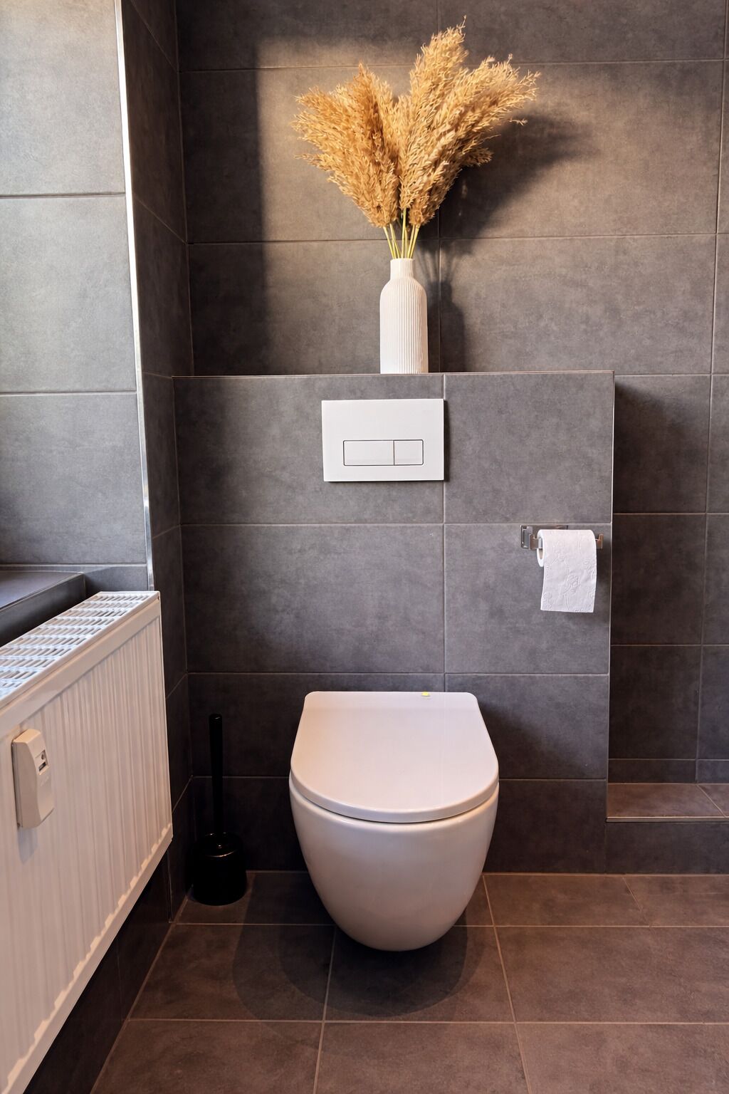 Une salle de bain avec des toilettes blanches, un radiateur, un interrupteur fixé au mur et un vase contenant des plantes séchées.