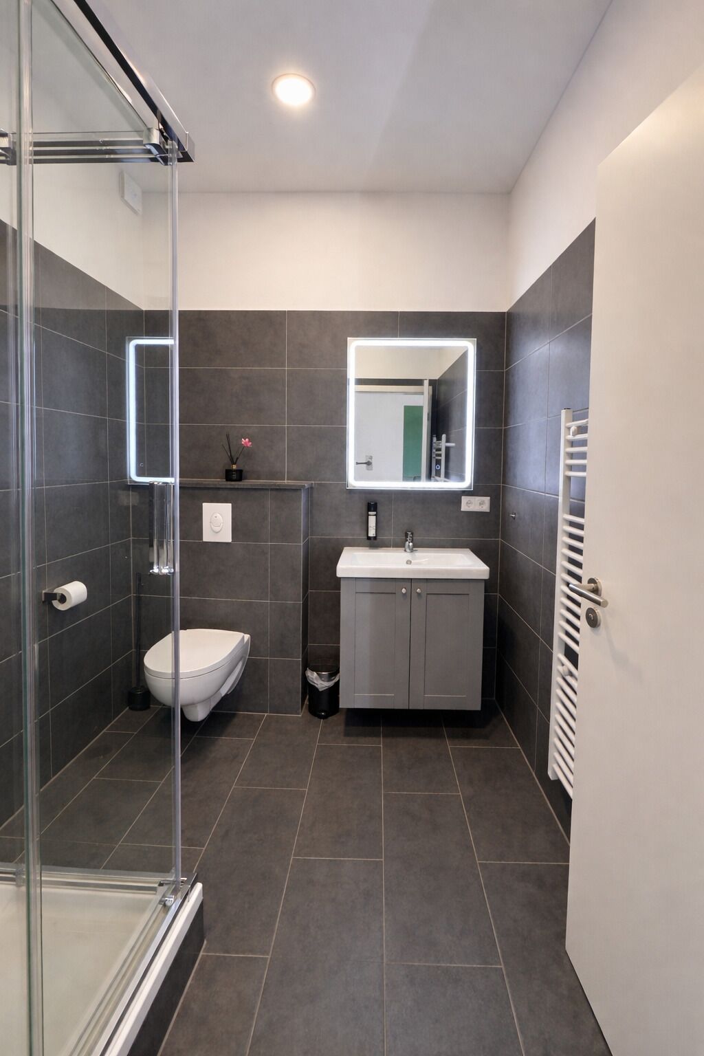 Un baño moderno con cabina de ducha de vidrio, inodoro blanco, lavamanos con espejo y un perchero para toallas.