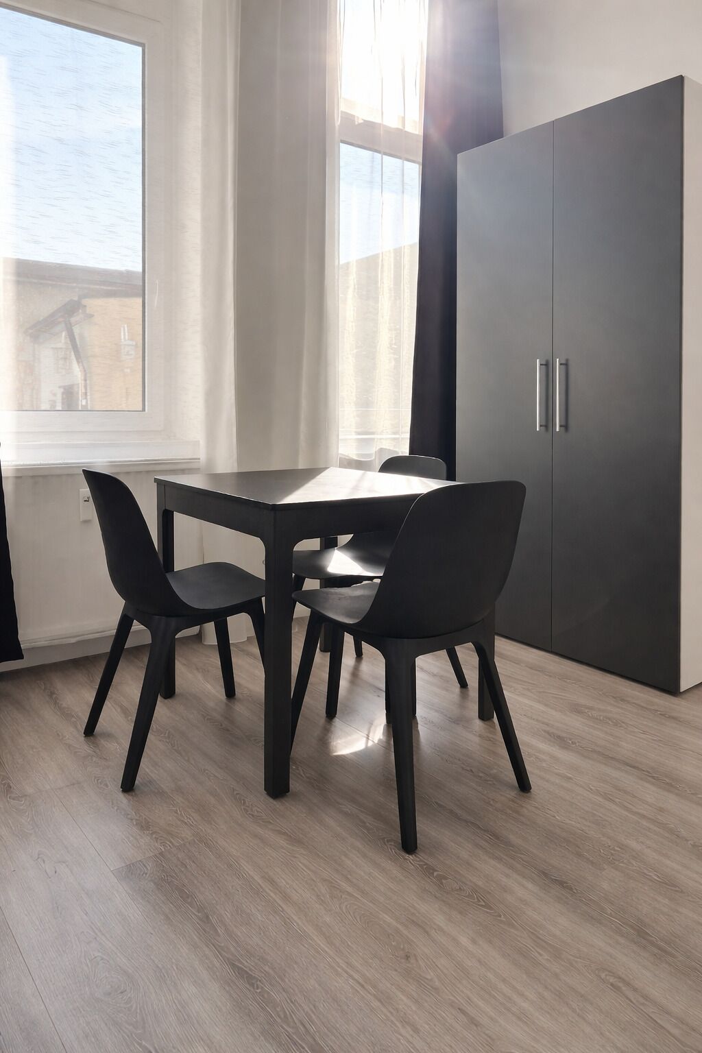 Un comedor moderno con una mesa, sillas, un armario oscuro y una ventana con cortinas.