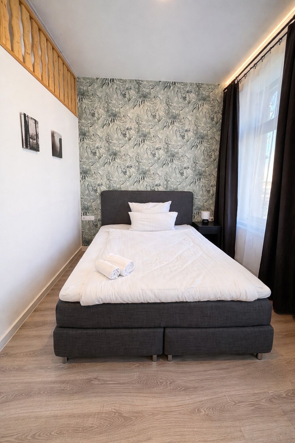 Een slaapkamer met een bed, een nachtkastje, een muur met een bladmotief en een raam met gordijnen.