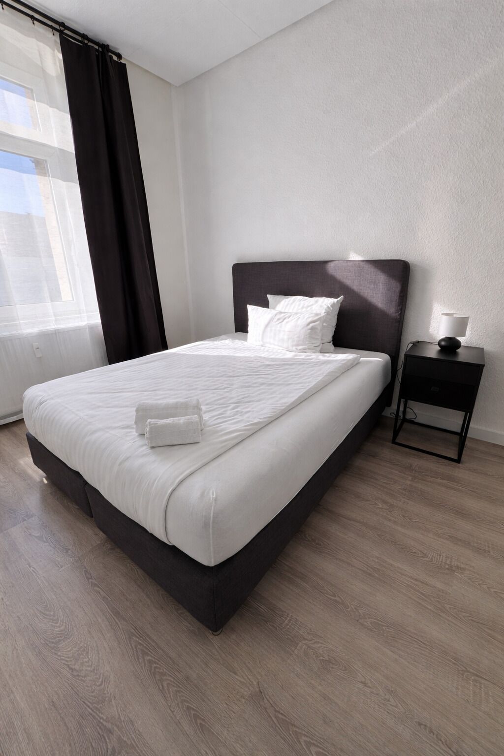 Una habitación de hotel moderna con una cama grande, una mesita de noche con lámpara y un ventanal con cortinas.