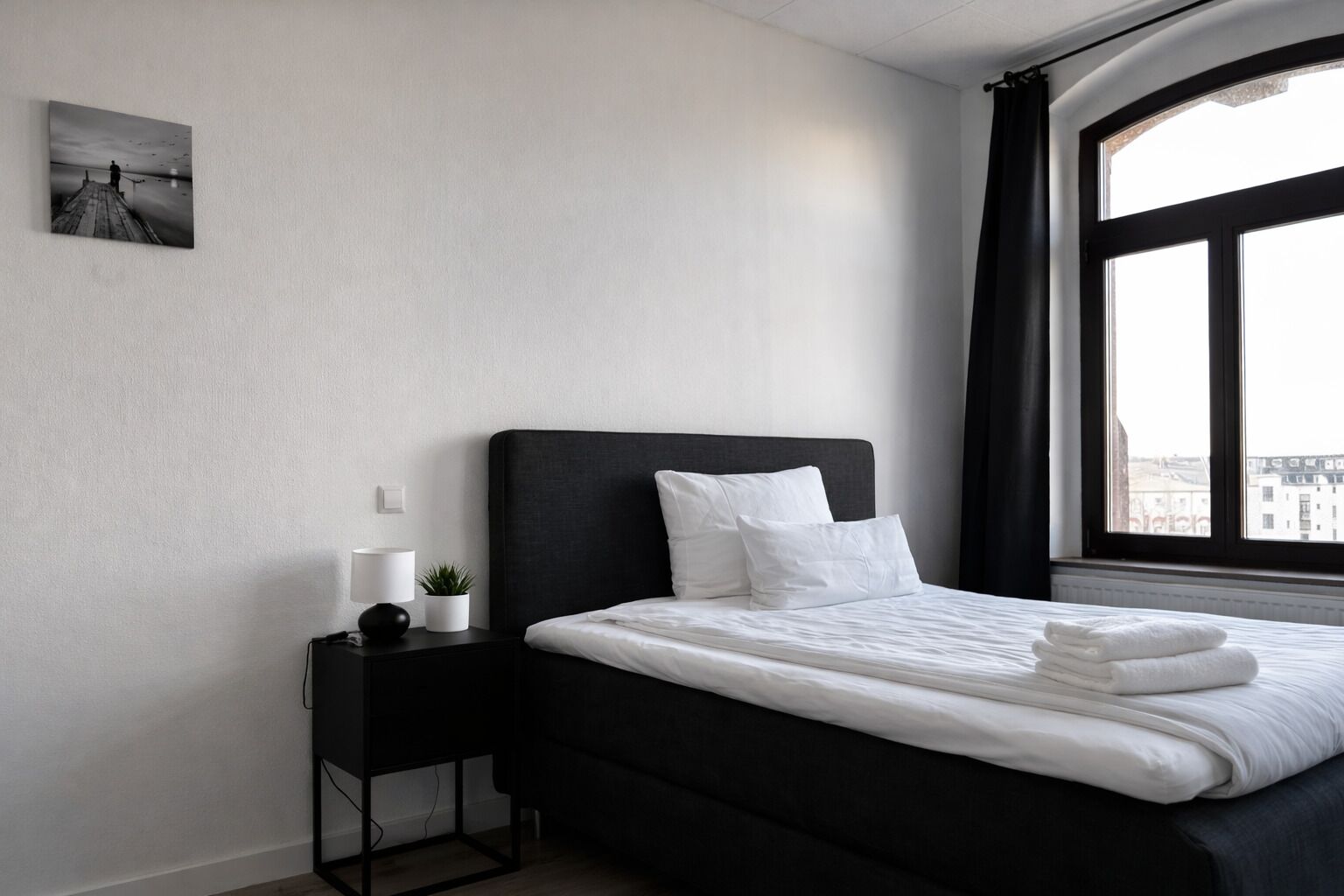 Ein modernes Hotelzimmer mit einem großen Bett, einem Nachttisch mit Lampe, einer kleinen Pflanze und Blick auf Gebäude durch ein Fenster.