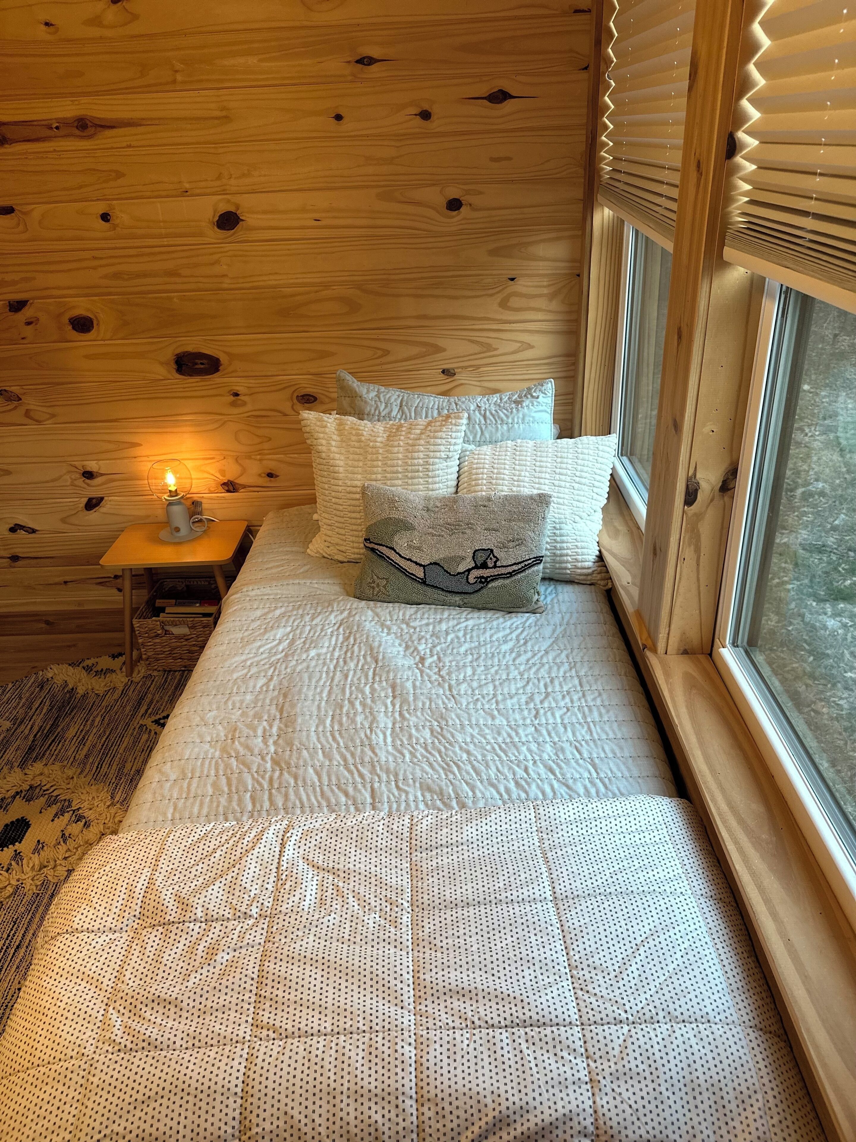 A wooden room with a bed, pillows, a nightstand with a lamp, and a window with blinds.