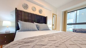 A hotel room with a large bed, a nightstand with a lamp, a window with a view of buildings, and decorative wall art. - San Isidro financial district stay (404) (Lima)
