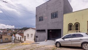 A modern building with a garage, a car parked in front, and a view of a residential area in the background. - Apartamento Para 6 h Spedes com Wi-fi e Garagem (Para de Minas)
