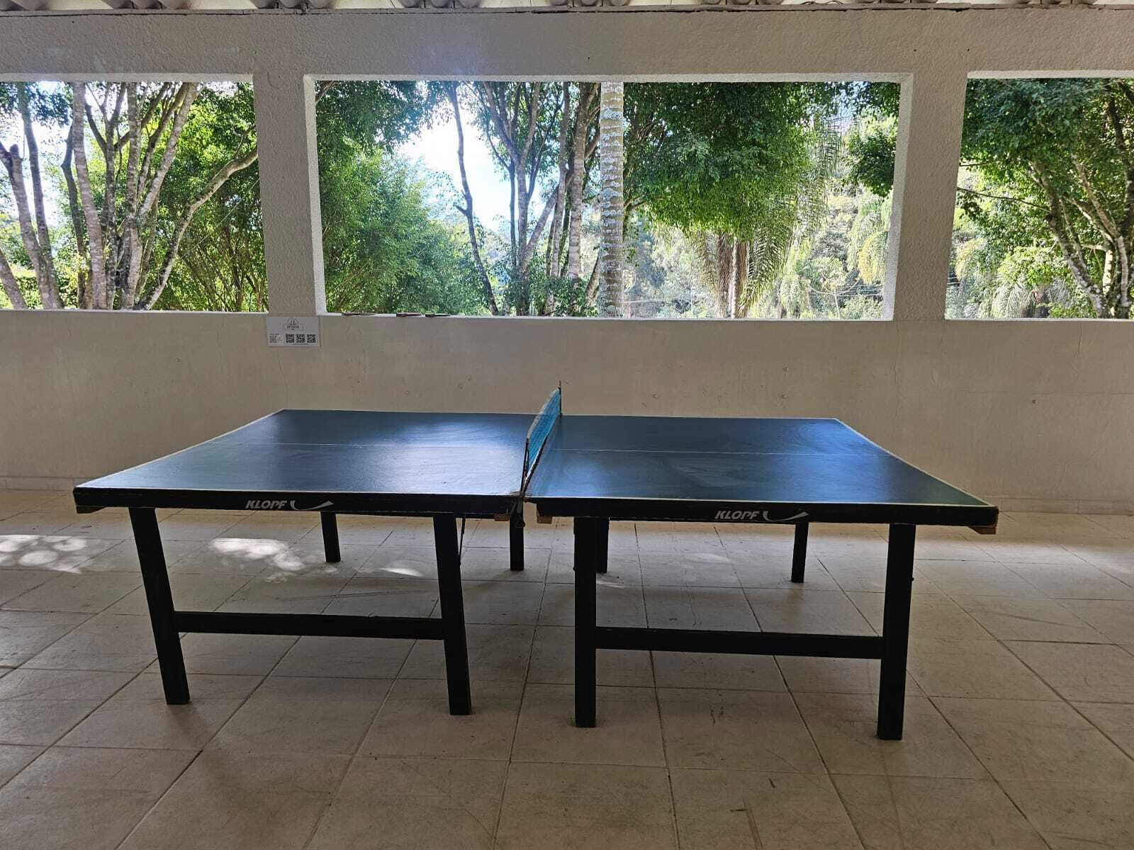 Uma mesa de pingue-pongue em um ambiente com piso ladrilhado e grandes janelas.