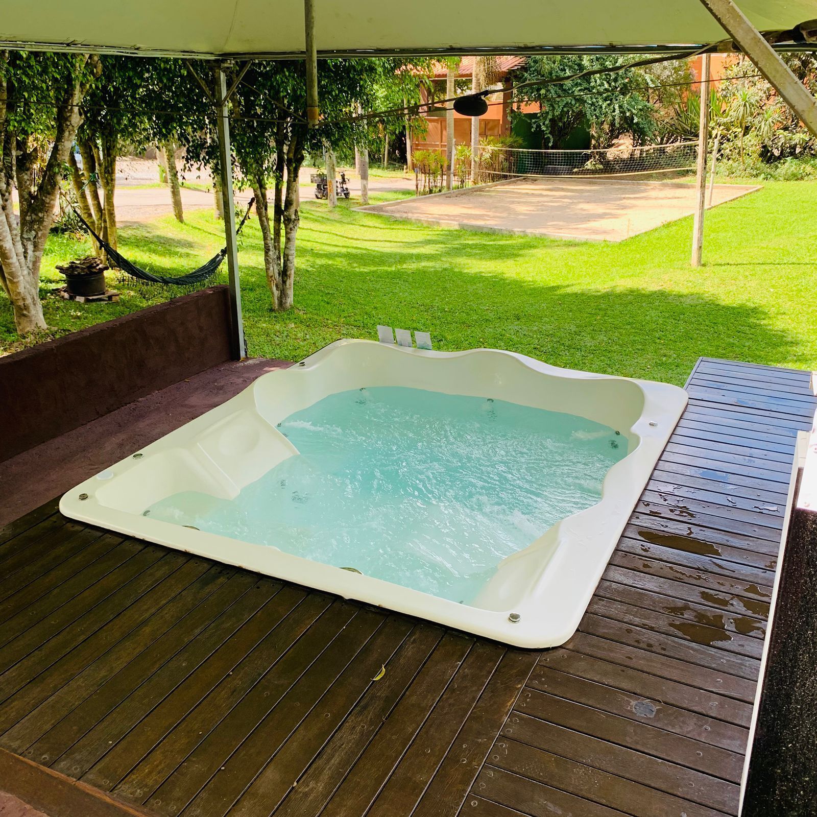 Un jacuzzi bajo una carpa, con un patio cubierto de césped y árboles al fondo.