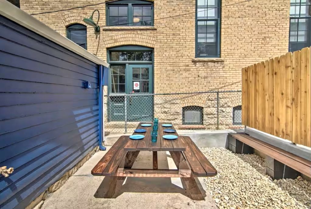 A wooden picnic table with plates and a water bottle, set against a brick building with a stone fireplace and a wooden fence.