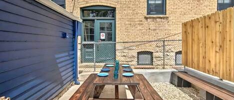 A wooden picnic table with plates and a water bottle, set against a brick building with a stone fireplace and a wooden fence.