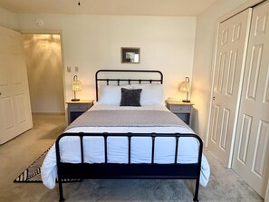 A bedroom with a black metal bed frame, white bedding, two bedside tables with lamps, a framed picture on the wall, and a carpeted floor. - Birdie House on Balboa (Hot Springs Village)