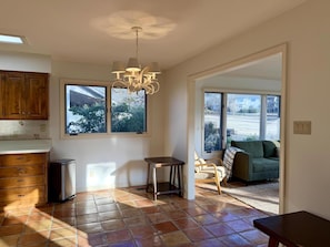 Cocina amplia con armarios de madera, refrigerador de acero inoxidable, una mesa de comedor y una araña. Amplias ventanas ofrecen luz natural y vistas al exterior.