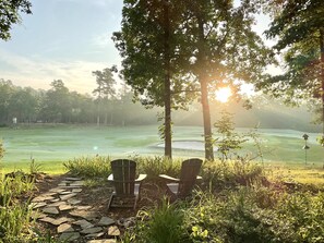 A stone pathway leading to two Adirondack chairs overlooking a golf course with a misty background and a sunlit sky. - Birdie House on Balboa (Hot Springs Village)